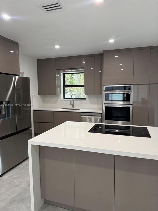 Modern kitchen featuring glossy taupe cabinetry, a large central island with a solid-surface waterfall countertop, a stainless steel double wall oven, and a built-in electric cooktop