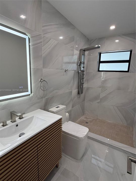 Bathroom featuring a fluted wood-finish vanity with a white basin, large-format tile wall cladding, an LED-backlit mirror, and a walk-in shower with a multi-jet panel system