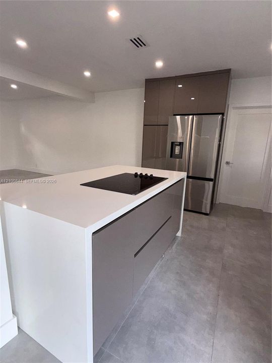 Kitchen island featuring a white stone countertop with an integrated electric cooktop and taupe cabinetry, stainless steel French-door refrigerator, and large-format tile flooring