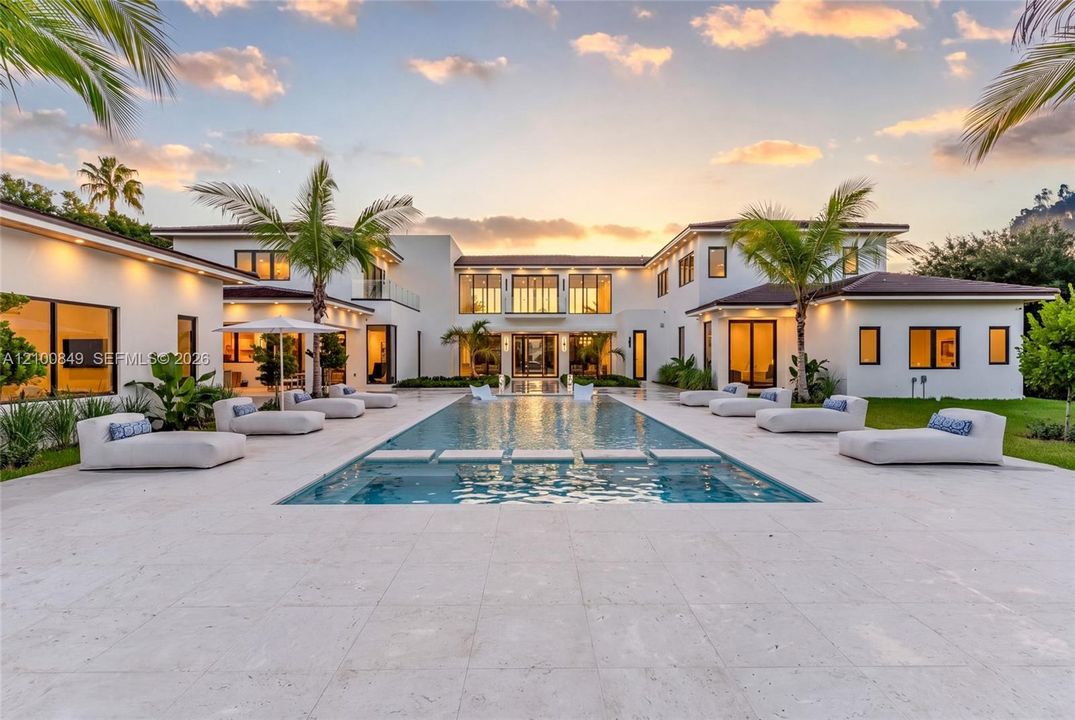 Contemporary estate exterior featuring a sprawling limestone pool deck, rectangular swimming pool with integrated stepping stones, multi-wing architectural design, and extensive glass-paneled windows