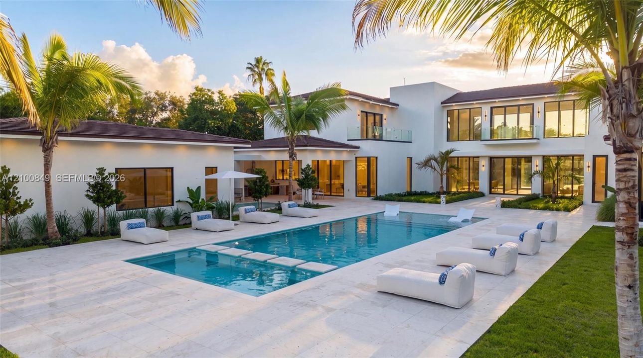 Custom pool with built-in stepping stones, expansive stone-paved patio, two-story white stucco exterior, multiple glass balcony railings, and lush perimeter landscaping