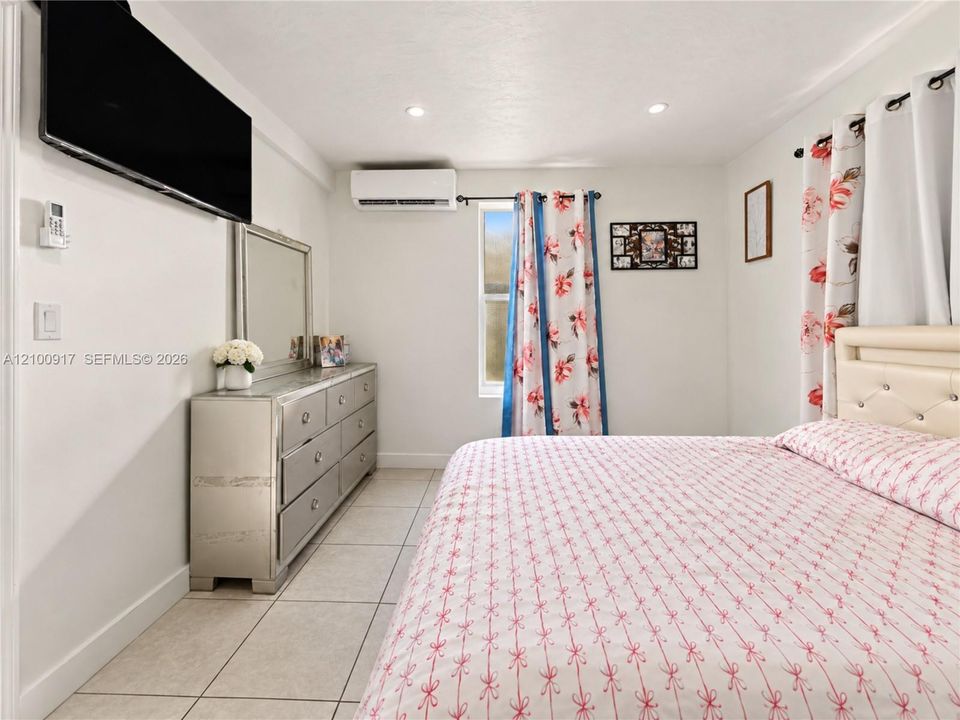 Recessed lighting, wall-mounted air conditioning unit, light-toned tile flooring, and a window with curtain rod