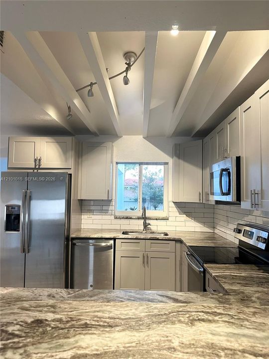 Kitchen featuring neutral shaker-style cabinetry, patterned stone-surface countertops, white subway tile backsplash, stainless steel appliances, and exposed ceiling beams