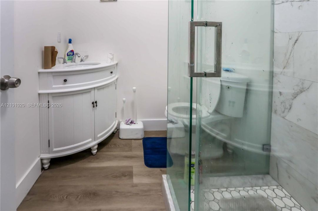 Curved white vanity with a ceramic basin, wood-finish flooring, glass-enclosed shower, and stone-tile shower surround with mosaic tile flooring
