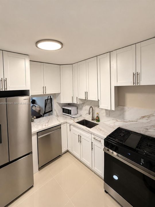 Kitchen featuring white Shaker-style cabinetry, stone-patterned countertops, stainless steel appliances, a deep basin under-mount sink with a high-arch faucet, and light-toned tile flooring