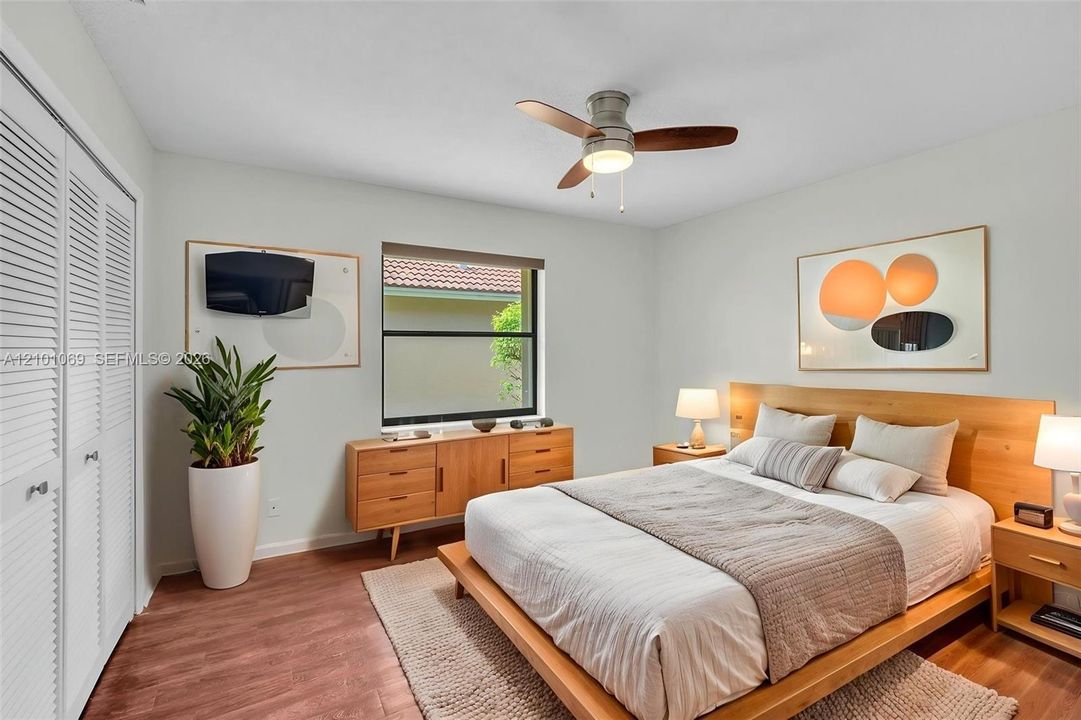 Bedroom featuring wood-finish flooring, white louvered closet doors, a central ceiling fan with integrated lighting, and a window with a neutral roller shade -virtually staged