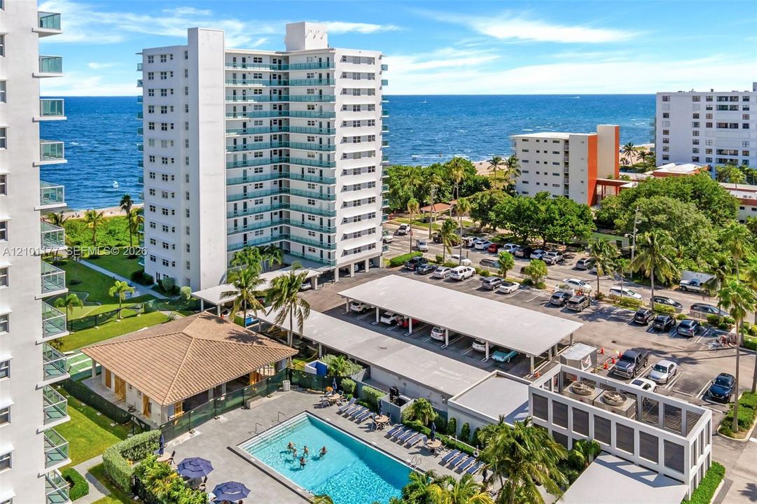 High-rise coastal property featuring an outdoor swimming pool, covered parking structures, tile-roofed amenity building, and established landscaping