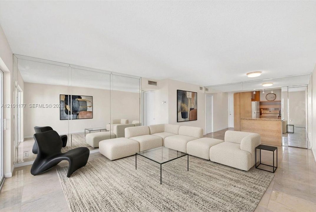 Open-concept living area featuring full-wall mirrors, light-toned stone-finish flooring, and a kitchen peninsula with cabinetry