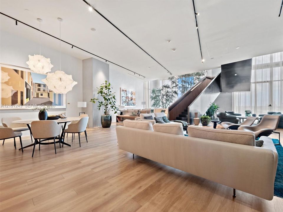 Open-concept main living area featuring light wood-finish flooring, floor-to-ceiling windows, sculptural statement pendant lighting, track lighting, and a floating architectural staircase with a metal railing