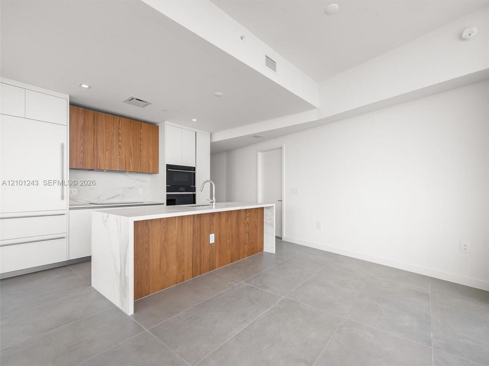 Kitchen featuring a central island with a solid surface waterfall countertop, two-tone cabinetry in white and wood-finish, stone-patterned backsplash, and large-format tile flooring