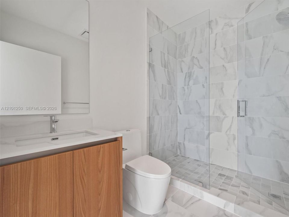 Bathroom featuring a vanity with a light-toned wood-finish cabinet and solid-surface countertop, a frameless glass-enclosed shower with large-format wall tile, and a wall-mounted mirror