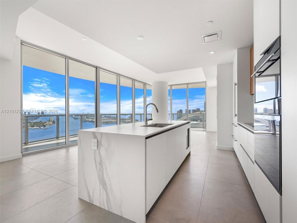 Kitchen featuring a central island with stone-finish waterfall edges, stainless steel under-mount sink, integrated appliances, floor-to-ceiling windows, and tile flooring