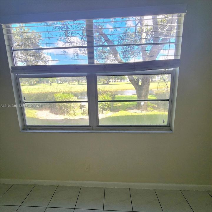Large dual-pane window featuring a horizontal blind system, ceramic tile flooring, and exterior landscape scenery including mature trees and a pond