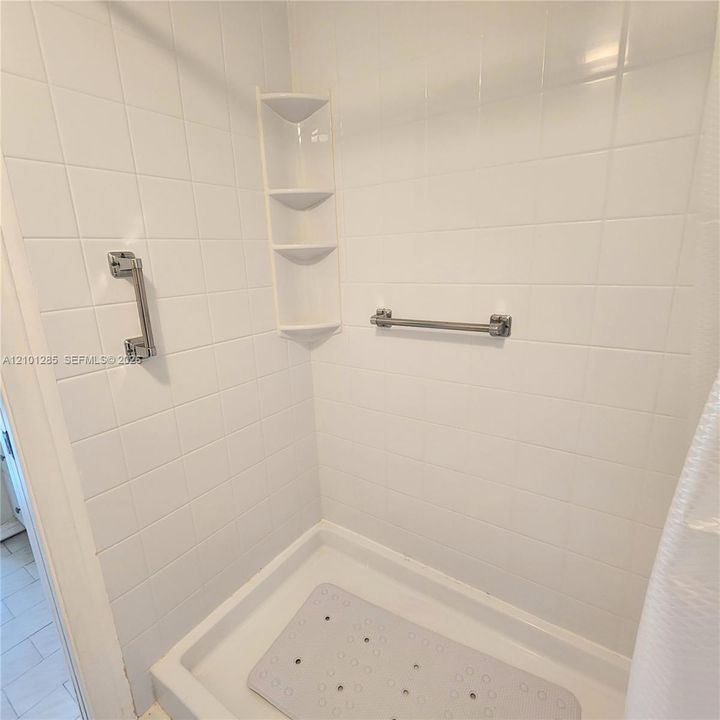 Shower stall featuring white square tile surround, built-in corner shelving, and dual stainless steel grab bars