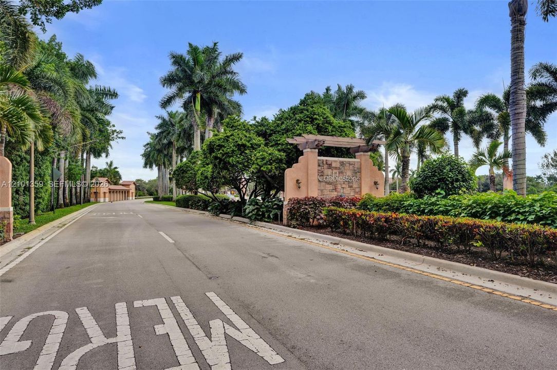 Stone-accented community monument signage featuring a wood-beam pergola, paved private roadway, and mature landscaping with established palm trees