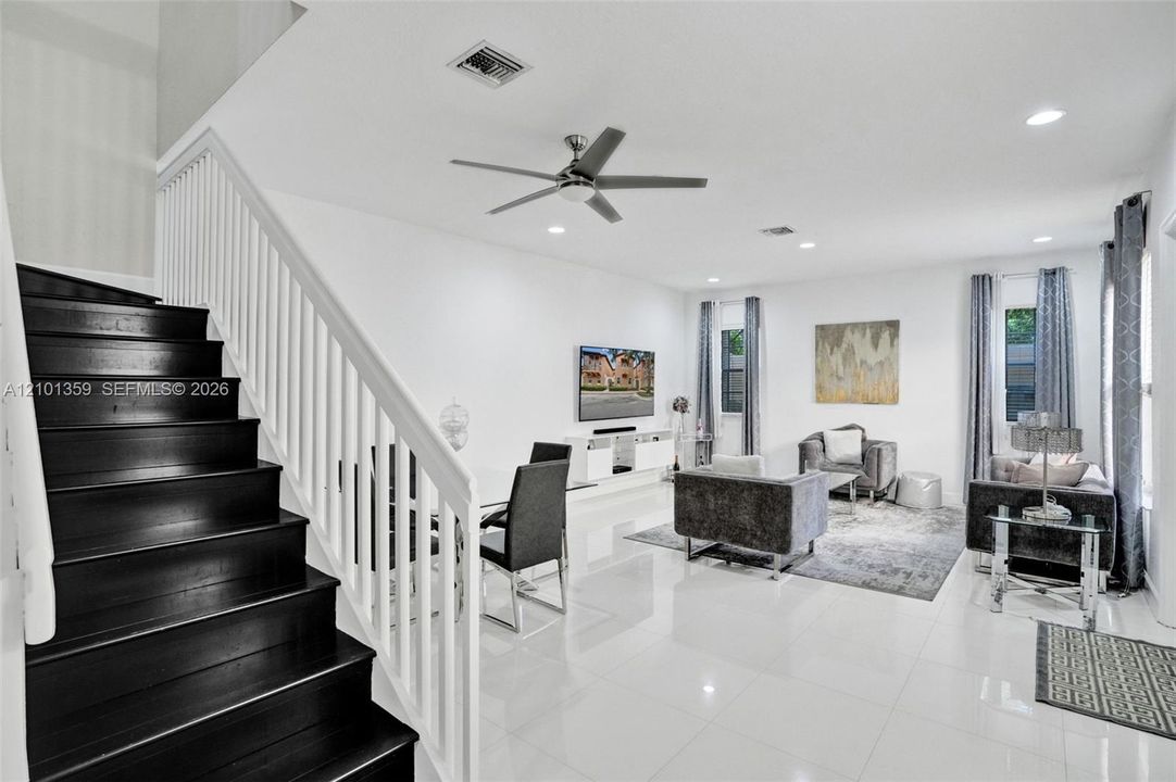 Open-concept interior featuring large-format tile flooring, a staircase with dark-finished treads and white handrails, recessed ceiling lighting, and a modern ceiling fan