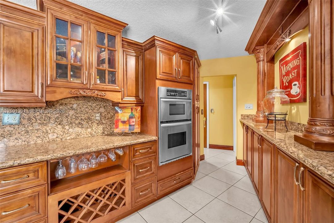 Custom wood cabinetry with glass-front accents, stainless steel double wall ovens, a built-in wine rack, and a stone-finish backsplash with countertops