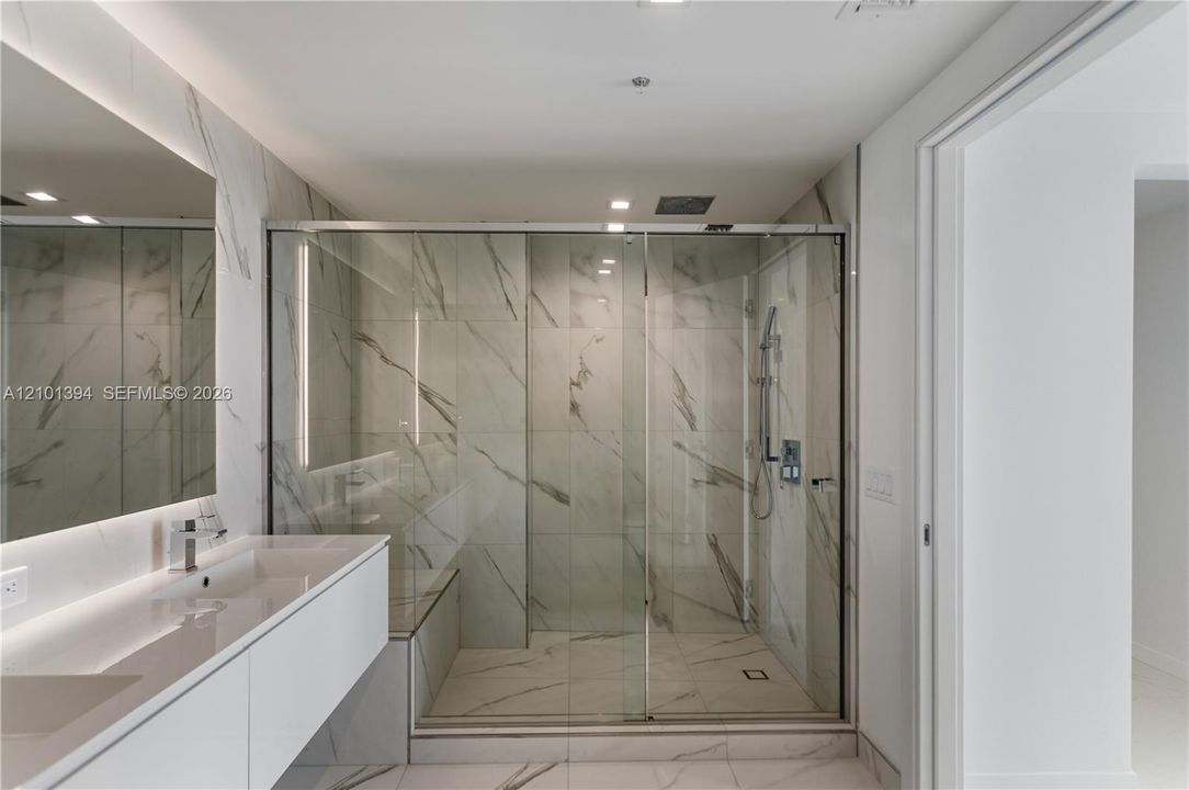 Floating white vanity with integrated dual sinks, large frameless wall-mounted mirror, and walk-in glass-enclosed shower featuring stone-finish wall and floor tile