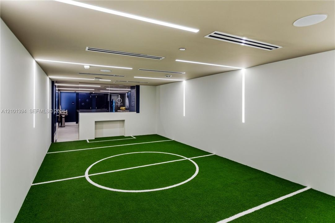 Indoor recreation area featuring synthetic turf flooring with integrated field markings, recessed linear ceiling lighting, vertical wall-mounted accent lighting, and a built-in counter