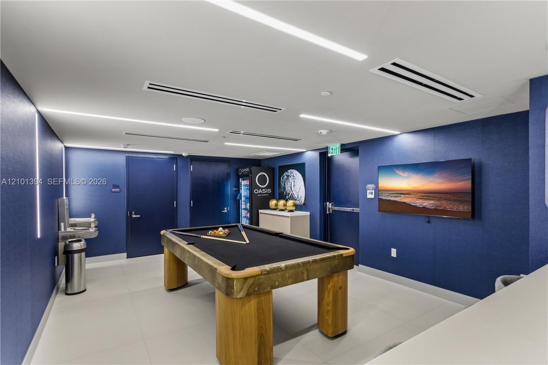 Rec room featuring recessed linear ceiling lighting, large-format tile flooring, integrated wall-mounted television, and blue textured wall surfacing