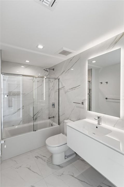 Floating vanity with integrated sink, backlit wall mirror, glass-enclosed shower and tub combination, and stone-finish floor and wall tile