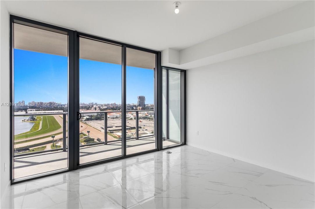 Floor-to-ceiling glass sliding doors, stone-finish tile flooring, and a private balcony with metal railings, featuring a cityscape and water horizon backdrop