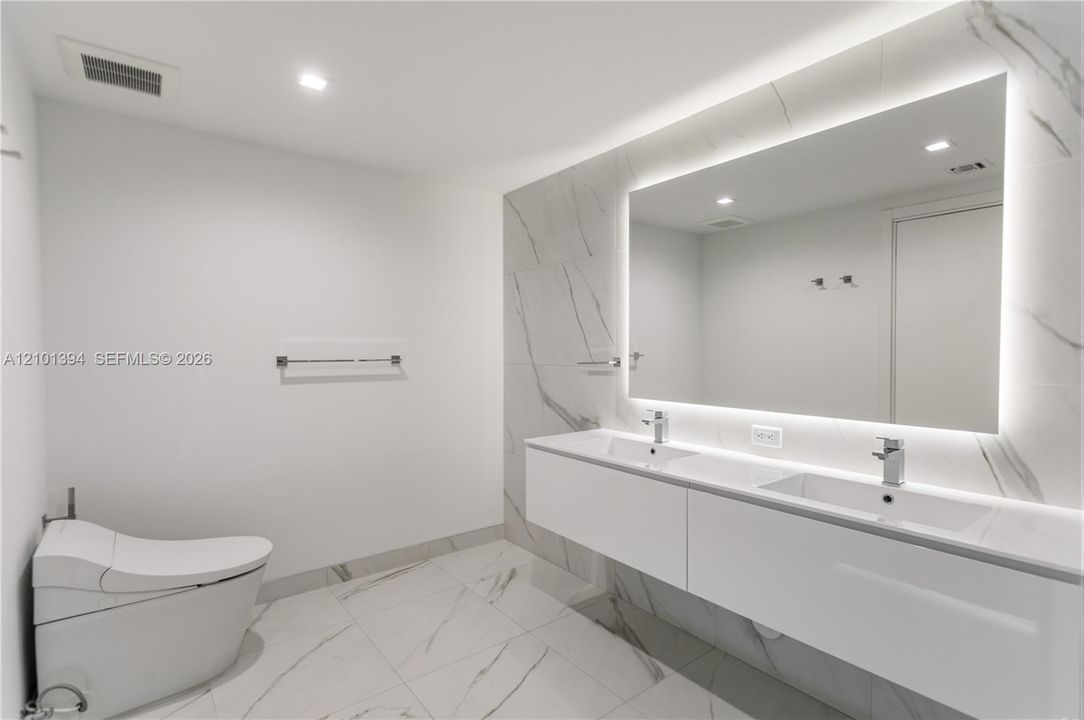 Modern floating dual-sink vanity with a solid surface countertop, large backlit mirror, and floor-to-ceiling stone-finish tile wall featuring a wall-mounted toilet