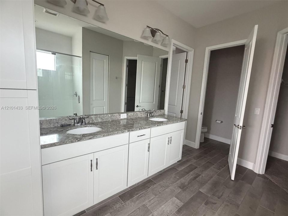 Dual-sink vanity with stone-finish countertop, white cabinetry, wall-to-wall mirror, and grey plank-style flooring