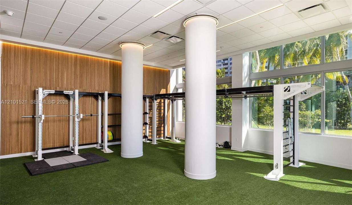 Fitness space featuring floor-to-ceiling windows, wood-paneled walls, synthetic turf flooring, recessed ceiling lighting, and structural cylindrical columns