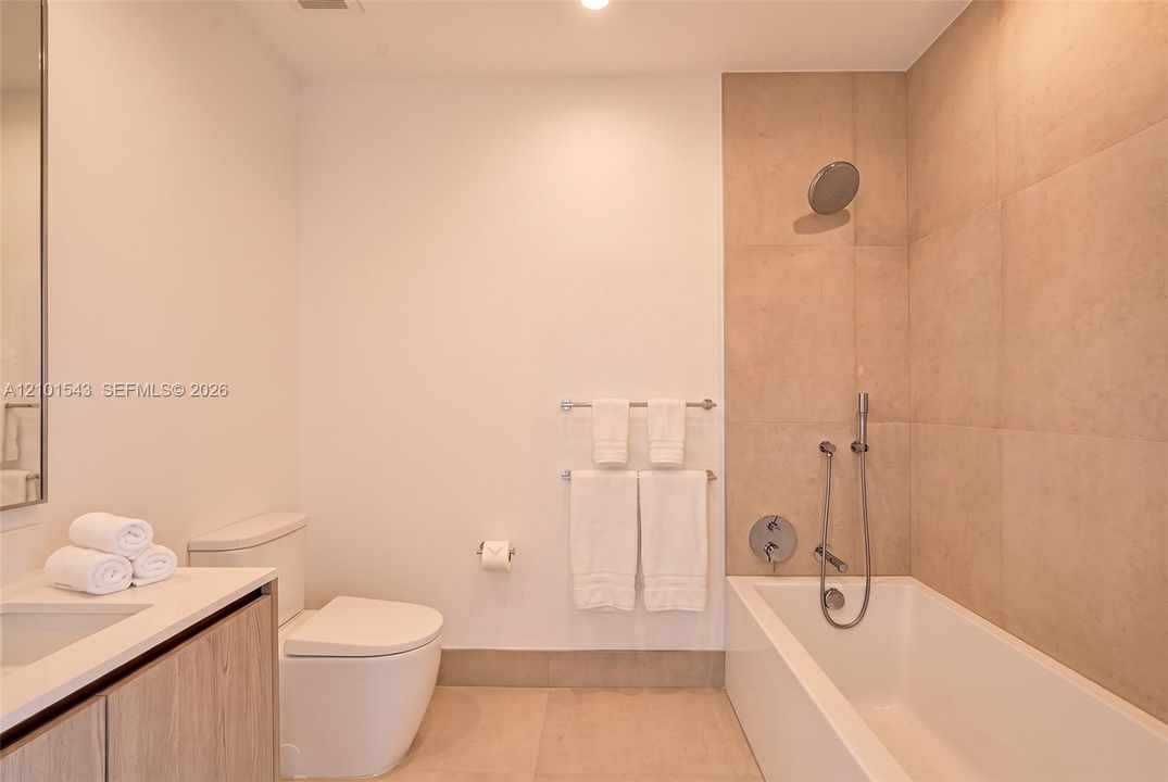 Bathroom featuring a wood-finish vanity, large-format neutral wall tile, integrated tub and shower combination with handheld fixture, and recessed lighting