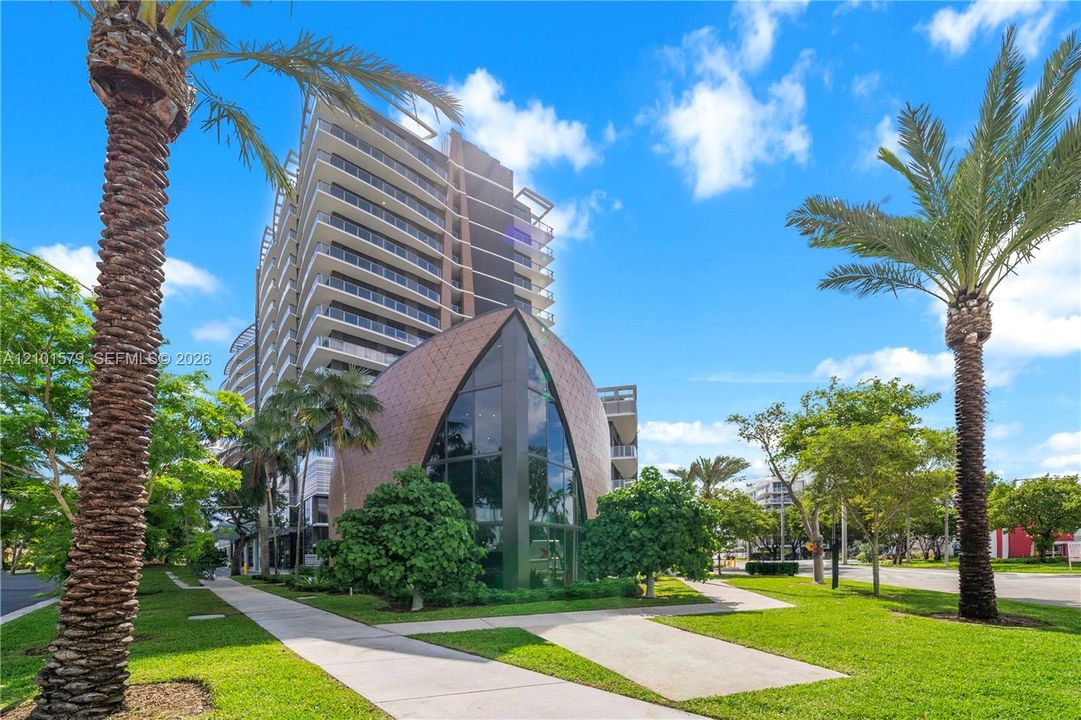 Multi-story architectural exterior featuring a curved metallic-toned facade, floor-to-ceiling glass paneling, tiered balconies, and manicured grounds with palm trees
