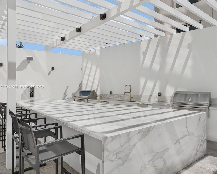 Outdoor kitchen featuring a large waterfall-edge countertop, stainless steel grill, undermount sink with a gooseneck faucet, and a white overhead pergola
