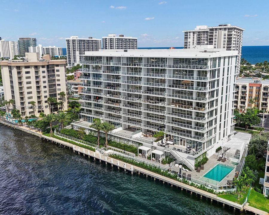 Waterfront mid-rise building featuring extensive private balconies, a ground-level swimming pool, manicured landscaping, and a direct bulkhead seawall