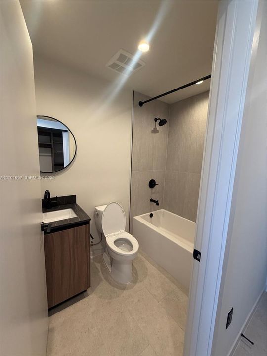 Bathroom featuring a wood-finish vanity with a dark stone countertop, circular mirror, and matte black hardware including a shower-tub faucet assembly with full-height tile surround