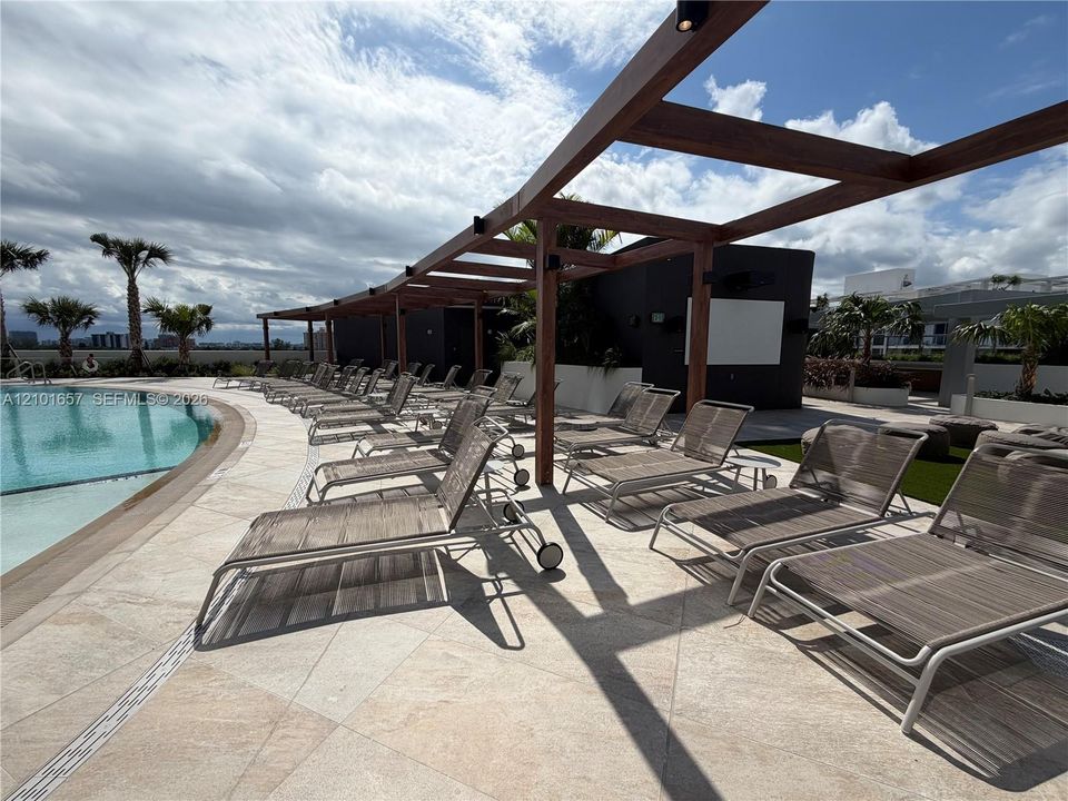 Curved swimming pool with stone-tiled decking and an overhead wooden pergola structure