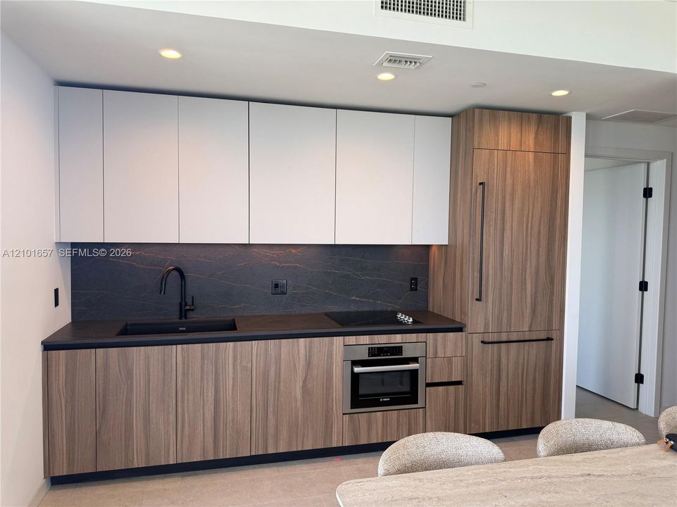 Modern kitchen featuring two-tone cabinetry with wood-finish lower cabinets, matte white upper cabinets, integrated refrigerator, stone backsplash, and recessed lighting