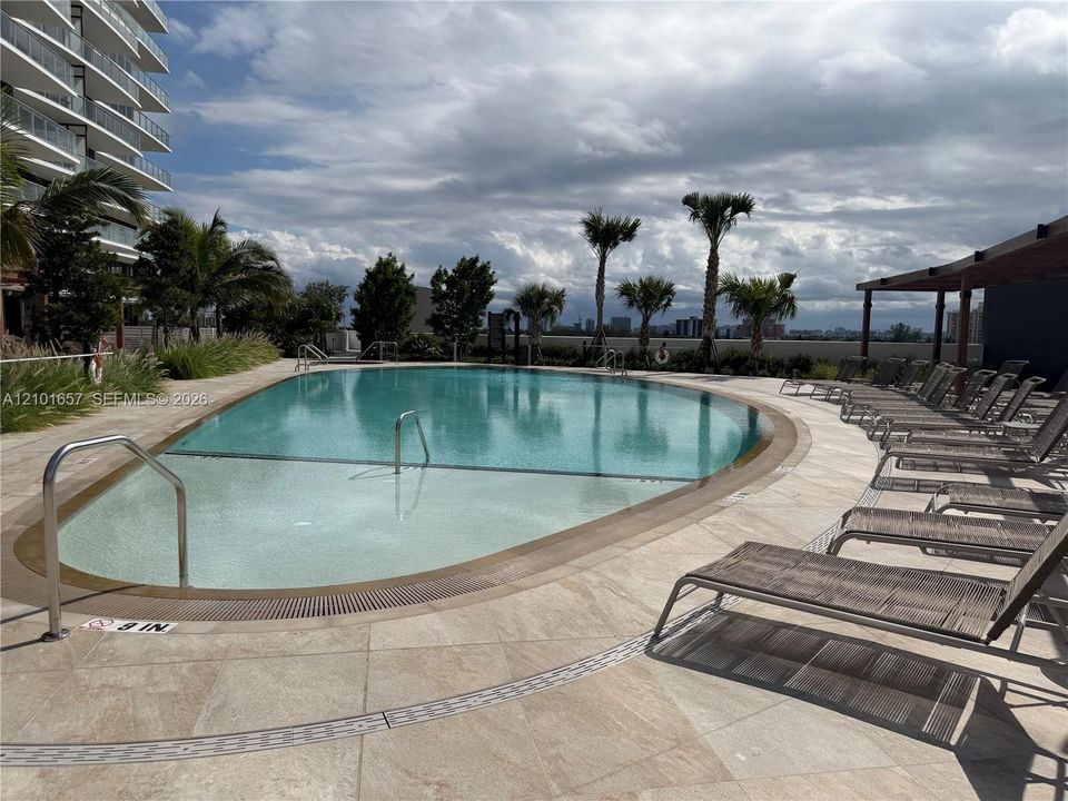 Curved swimming pool featuring a shallow entry shelf, tile decking, metal handrails, and a covered pergola