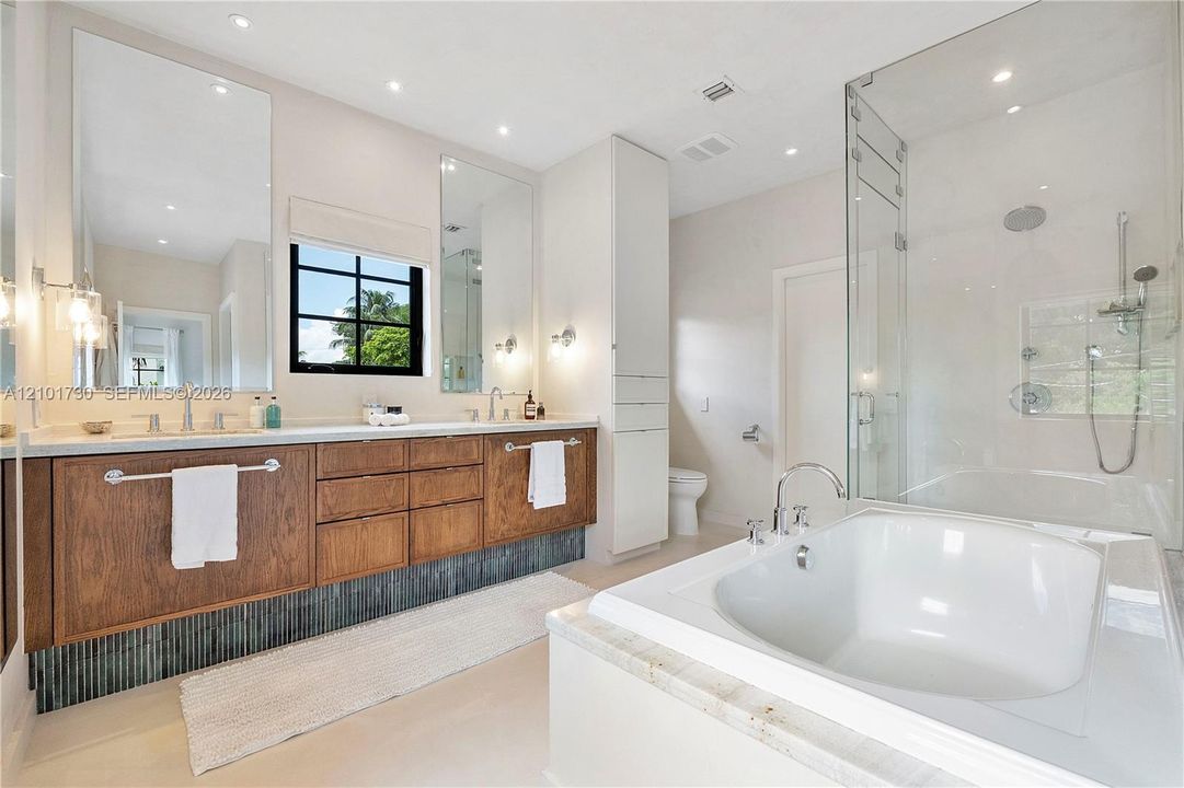 Bathroom featuring a dual-sink floating vanity with wood cabinetry, a glass-enclosed shower, a deep soaking tub with stone surround, and tall floor-to-ceiling built-in storage