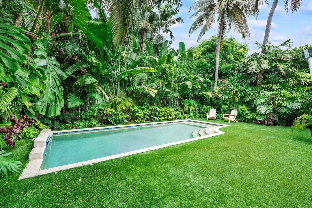 In-ground swimming pool featuring stone coping and built-in steps, surrounded by a lawn and dense tropical landscaping