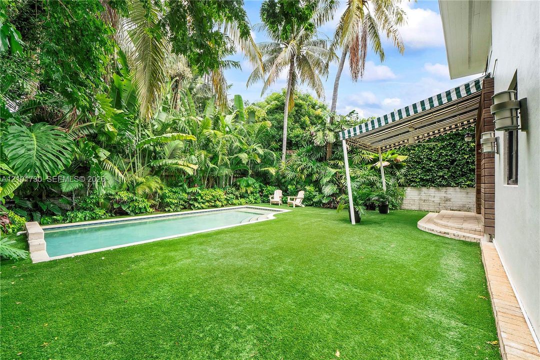 Private outdoor space featuring a rectangular swimming pool, manicured lawn, striped retractable awning, and mature tropical landscaping