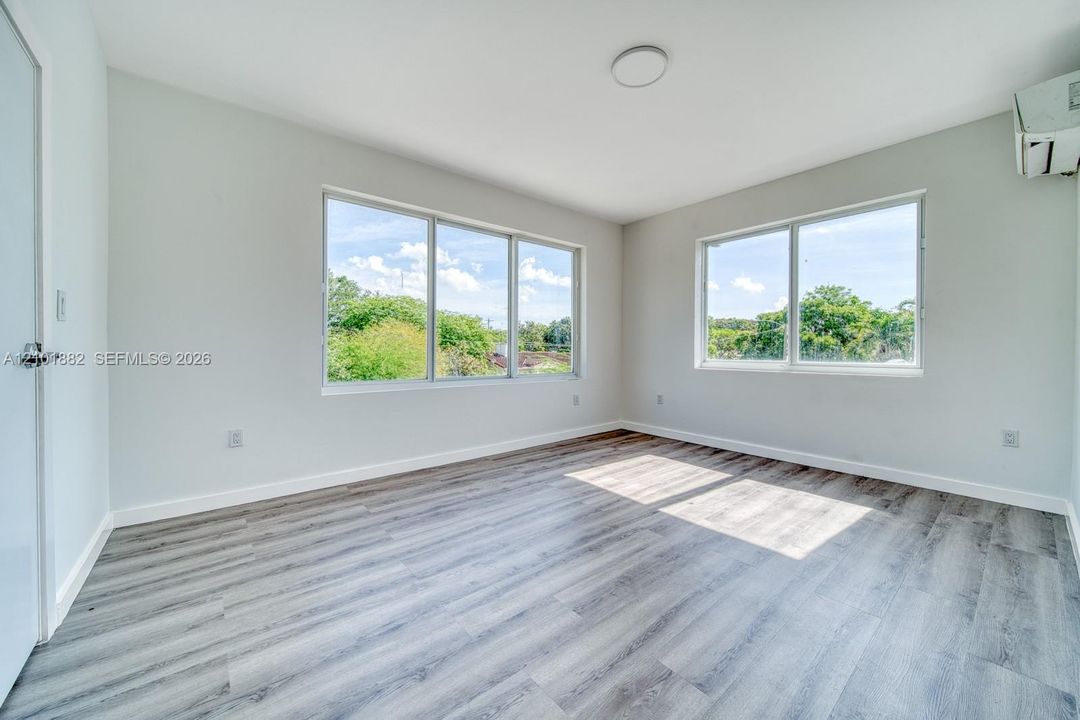 Neutral-toned interior featuring wood-finish flooring, oversized windows, and a wall-mounted climate control unit