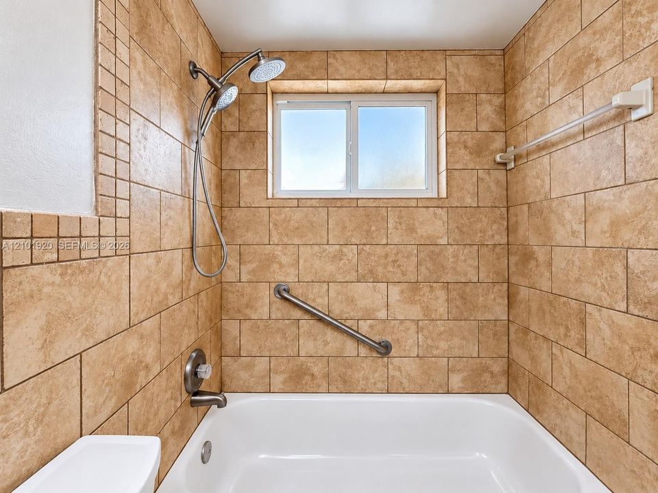 Full-height wall tile in a running bond pattern, white acrylic tub, adjustable dual-head shower fixture, built-in grab bar, and inset window