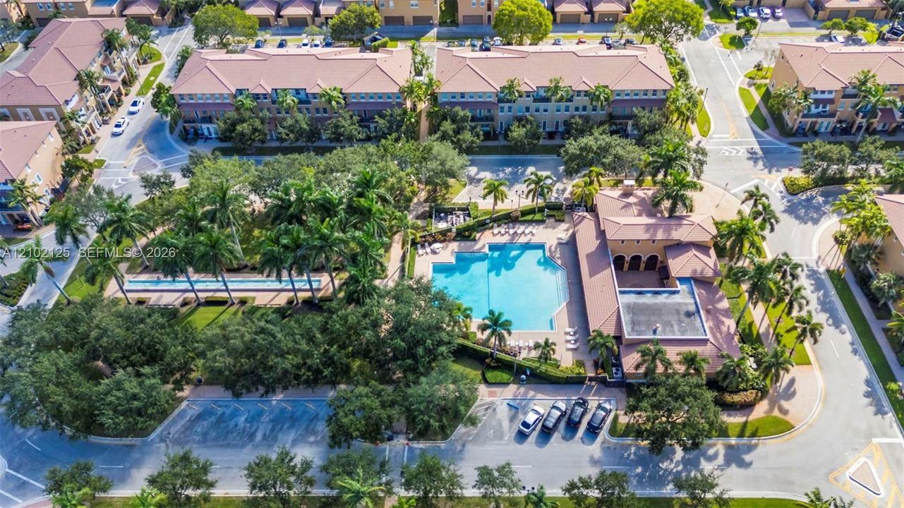 Aerial perspective of a community featuring a resort-style swimming pool, expansive sundeck, mature landscaping, palm tree clusters, and dedicated onsite parking