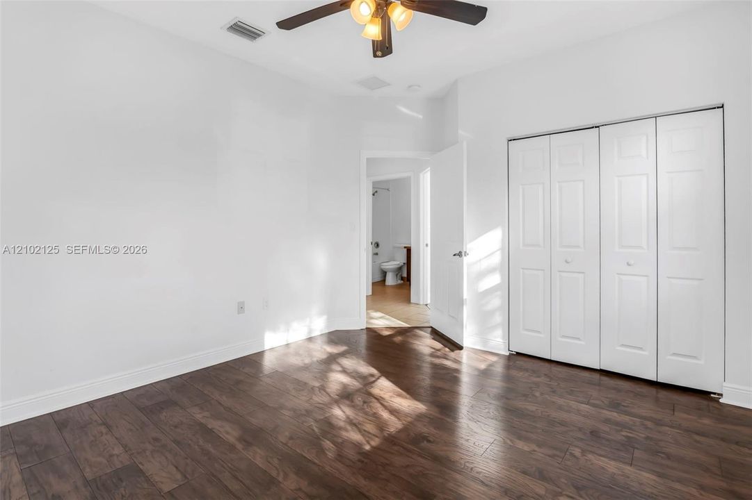 Spacious bedroom featuring rich wood-finish flooring, abundant natural light, ceiling fan with integrated lighting, and dual mirrored closet doors providing ample storage and reflective brightness throughout the room