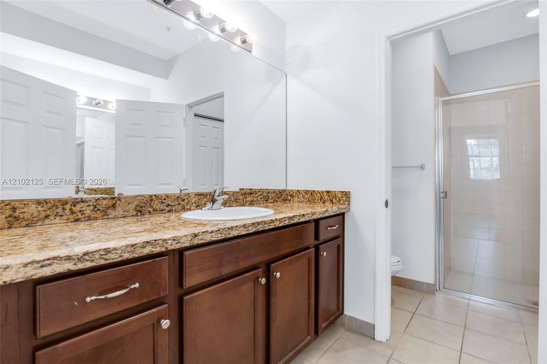 Bathroom vanity featuring a stone-composite countertop, undermount sink, wooden cabinetry, a full-width wall mirror, and a glass-enclosed shower stall with tiled flooring
