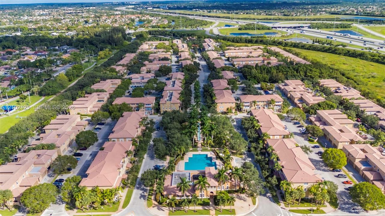 Aerial perspective of a multi-unit residential community featuring clay-tile roofing, a central swimming pool, paved surface parking, and mature palm tree landscaping