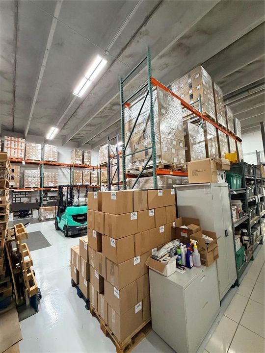 Concrete ceiling with exposed beams, fluorescent overhead lighting, warehouse racking system, concrete flooring, and filing cabinets