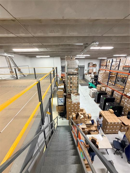 Concrete ceiling structure, industrial-grade fluorescent lighting, durable metal staircase with handrails, expansive warehouse flooring, and a multi-level storage system