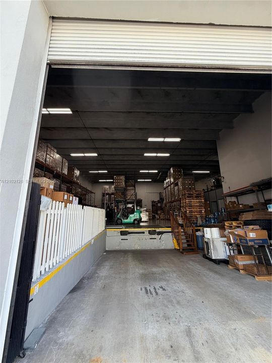 Overhead roll-up door entrance to a concrete floor warehouse with high ceilings, fluorescent lighting, and loading dock access
