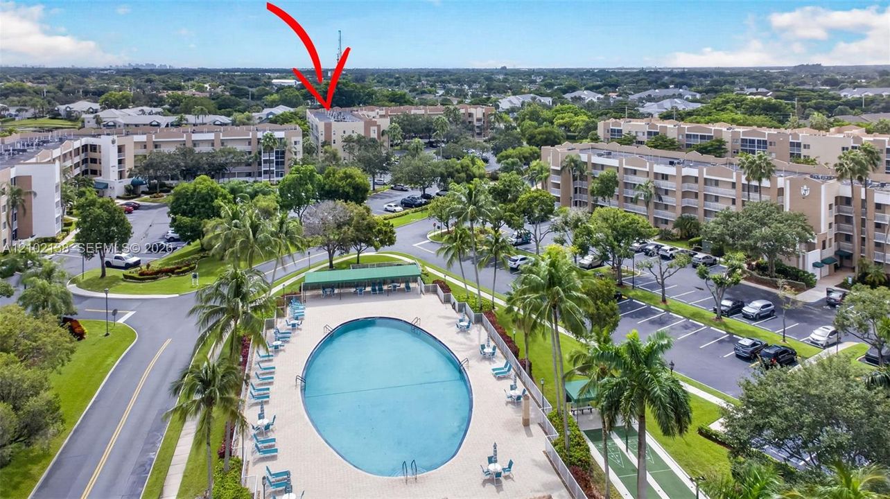 Aerial perspective of the condominium complex featuring a large swimming pool, paved parking areas, curated landscaping with palm trees, and designated shuffleboard courts